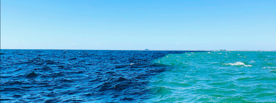 A wide seascape where two bodies of water with different colours meet along a visible line, deep blue on one side and bright turquoise on the other, without mixing.